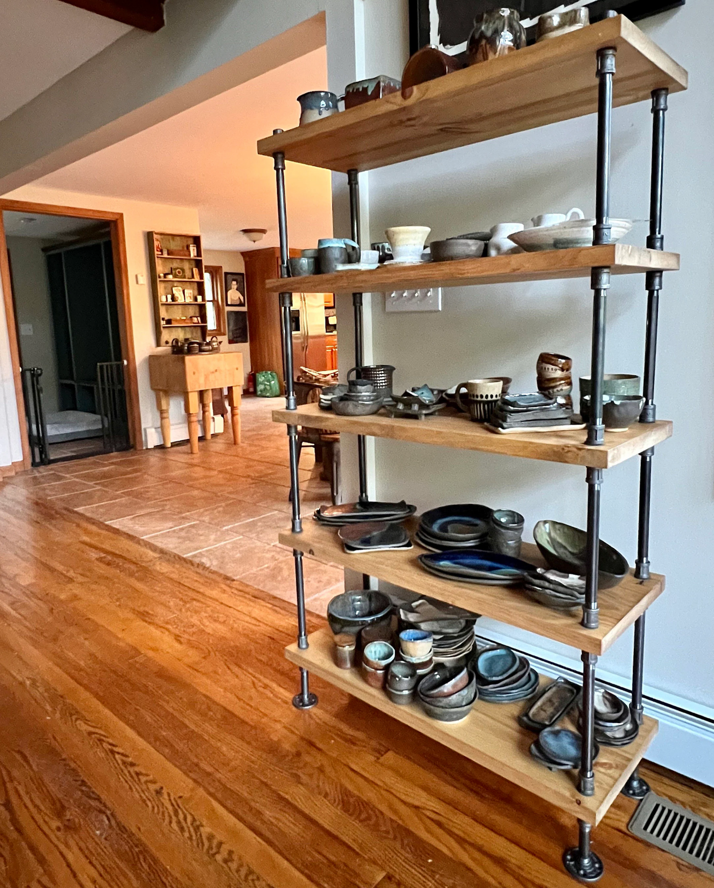 Handmade bookcase in industrial farmhouse style with natural wood shelves made of pine with a light yellow golden stain color applied. Bookcase is made with genuine black iron pipes for industrial vintage style. Bookcase sits in front of wall with handmade pottery such as dishes, cups, vases, glazed in many colors. Handmade bookcase by JT INDUSTRIAL DESIGNS in Texas.