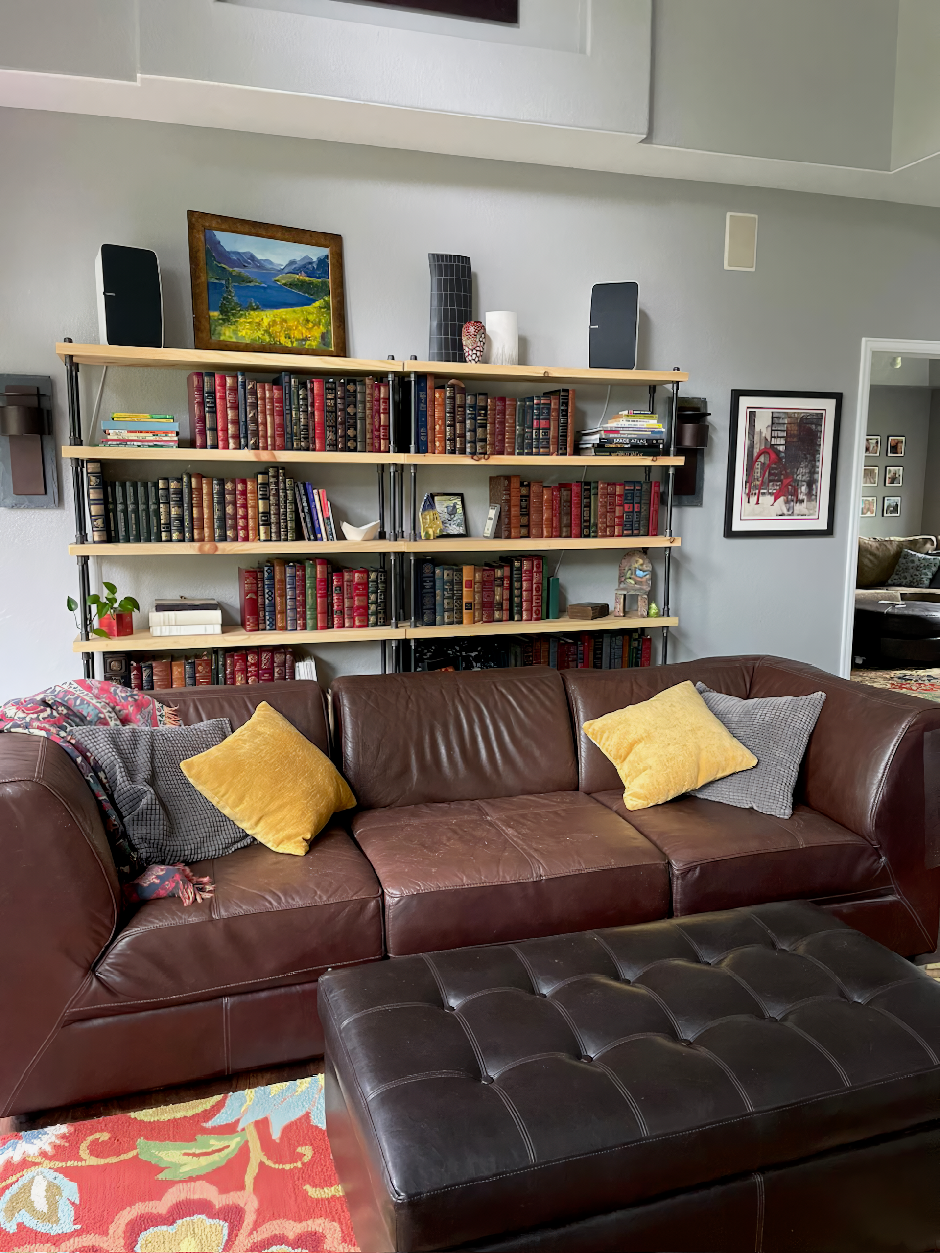 Idustrial famrhouse bookcase in large living room, book shelves are side by side for ease of storage and hold many tall heavy books. Bookcases sit behind a red leather couch that has cozy golden yellow pillows. Handmade wooden shelving units are the statement piece of the room design.