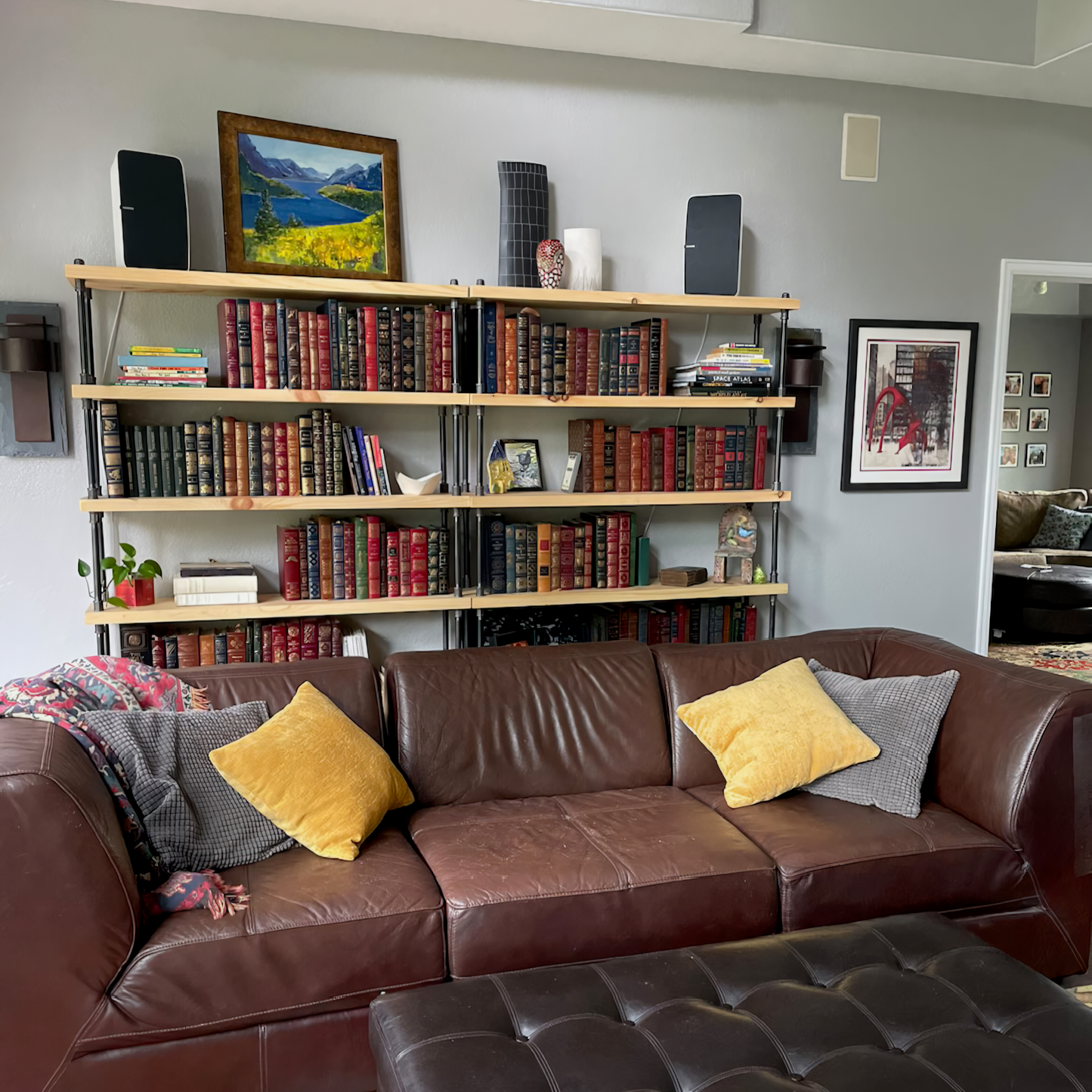 Industrial farmhouse bookcase in large living room, book shelves are side by side for ease of storage and hold many tall heavy books. Bookcases sit behind a red leather couch. Handmade wooden shelving units are the statement piece of the room design.