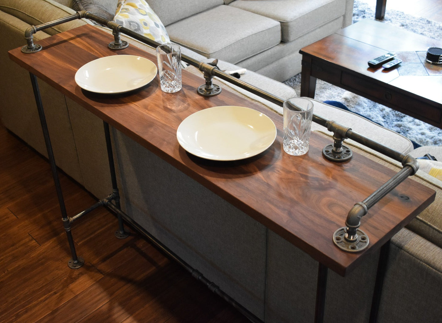 HARDWOOD WALNUT BAR TABLE BEHIND A GRAY SOFA, TWO WHITE DIINER PLATES AND TWO CRYSTAL GLASS CUPS SIT ON THE TABLE - JT INDUSTRIAL DESIGNS HANDMADE FURNITURE