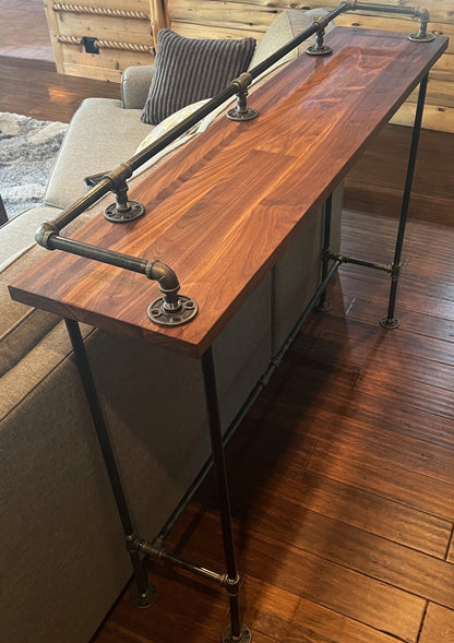 Hardwood walnut bar table top for kitchen or sofa table. Wood is solid with brown and blonde color tones. Genuine metal pipes and solid wood. Table is in living room behind gray sofa.
