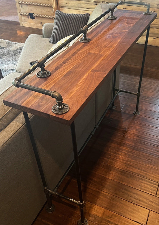 Hardwood walnut bar table top for kitchen or sofa table. Wood is solid with brown and blonde color tones. Genuine metal pipes and solid wood. Table is in living room behind gray sofa.