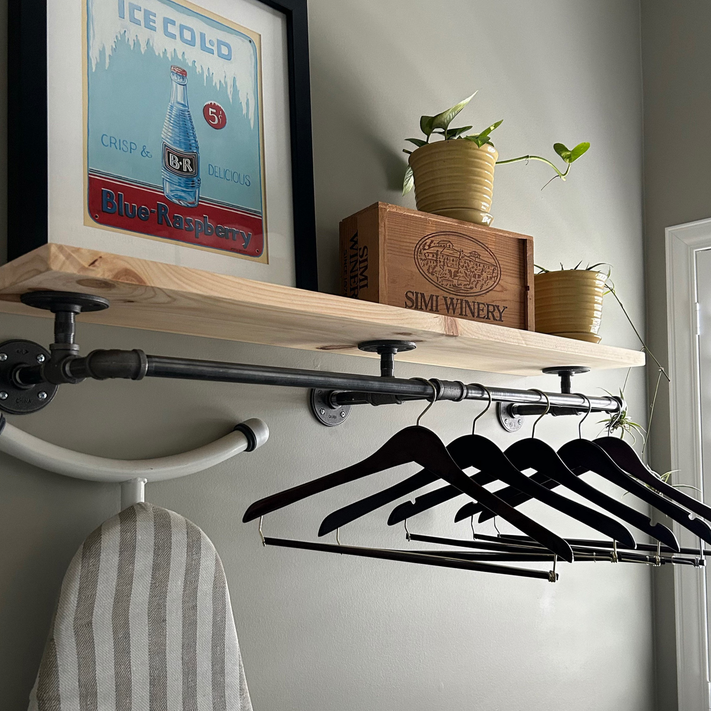JT Industrial Designs - hanger rack with wood shelf and industrial iron hanger bar. Natrual light yellow wood stain color on pine wood, metal is dark gray. Black hangers hang on the bar with painting and plant on shelf above.