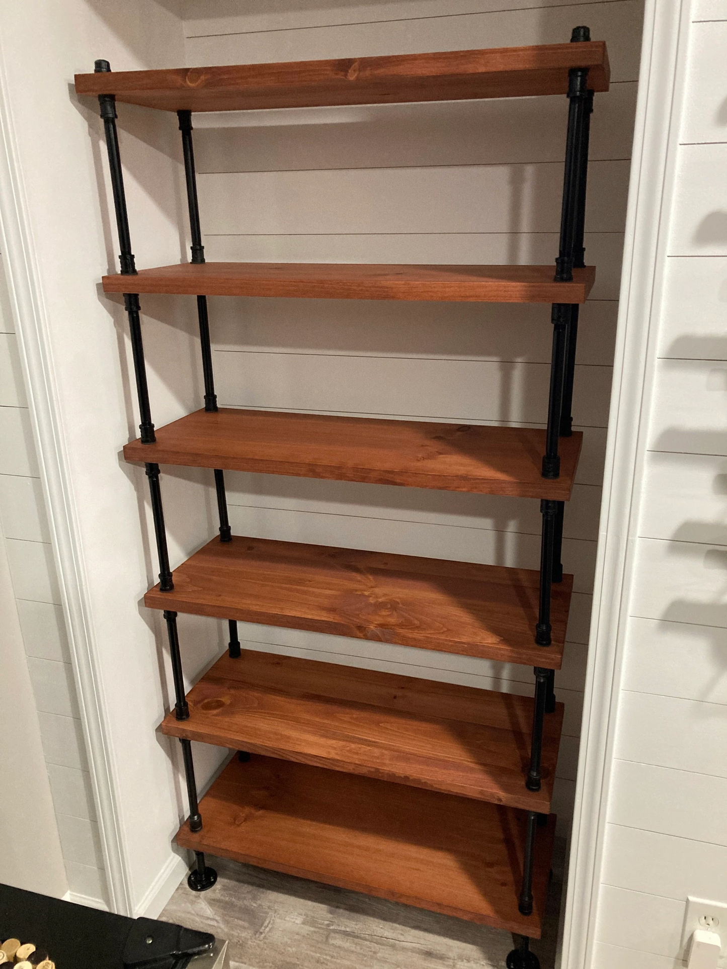 Bookcase sits in pantry kitchen nook with white walls. Bookcase is solid wood with balck iron pipe etagere frame. Shelves are solid pine wood finished with gunstock Minwax stain. Shelves are 36 inches length by 14 inches deep. Handmade bookcase by JT INDUSTRIAL DESIGNS in Texas.