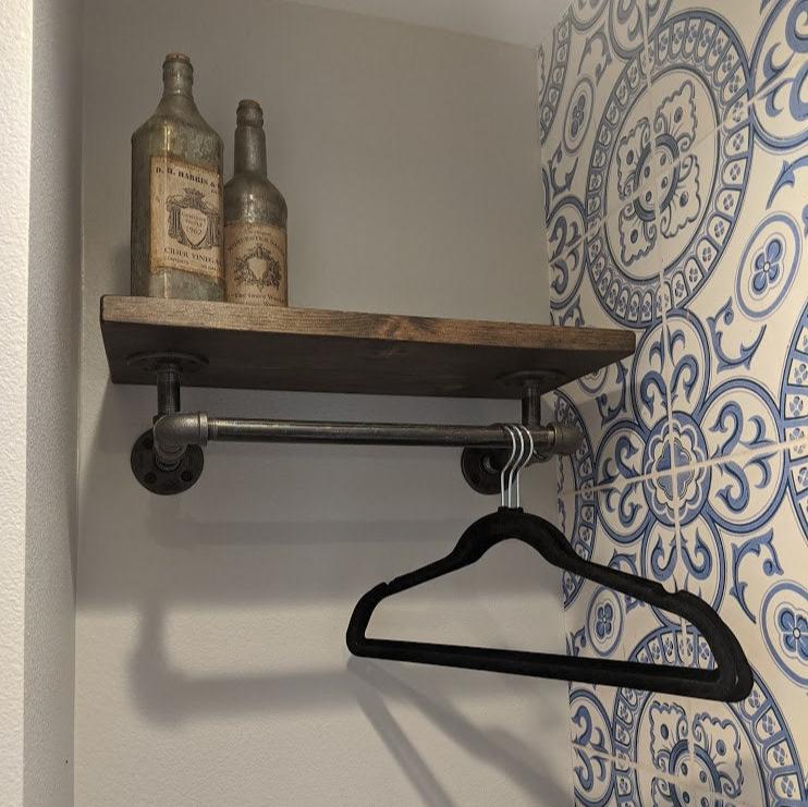 Industrial pipe with wood shelf hanger rack installed wall mounted with iron brackets, shelf is dark brown wood color. Hangers are hooked onto bar and decorative bottles on top of shelf. Ornate blue tile on the wall adjacent.