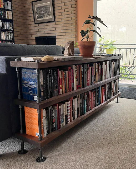 Industrial Bookcase unit with 3 shelves, 72 inches ling and 34 inches in hieght. Shelving unit is behind a dark blue sofa in a bright room with modern farmhouse decor style. Shelving unit has books lined up from end to end.